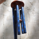 The Woodstock Dahlia Garden Wind Chime in front of a cement wall.
