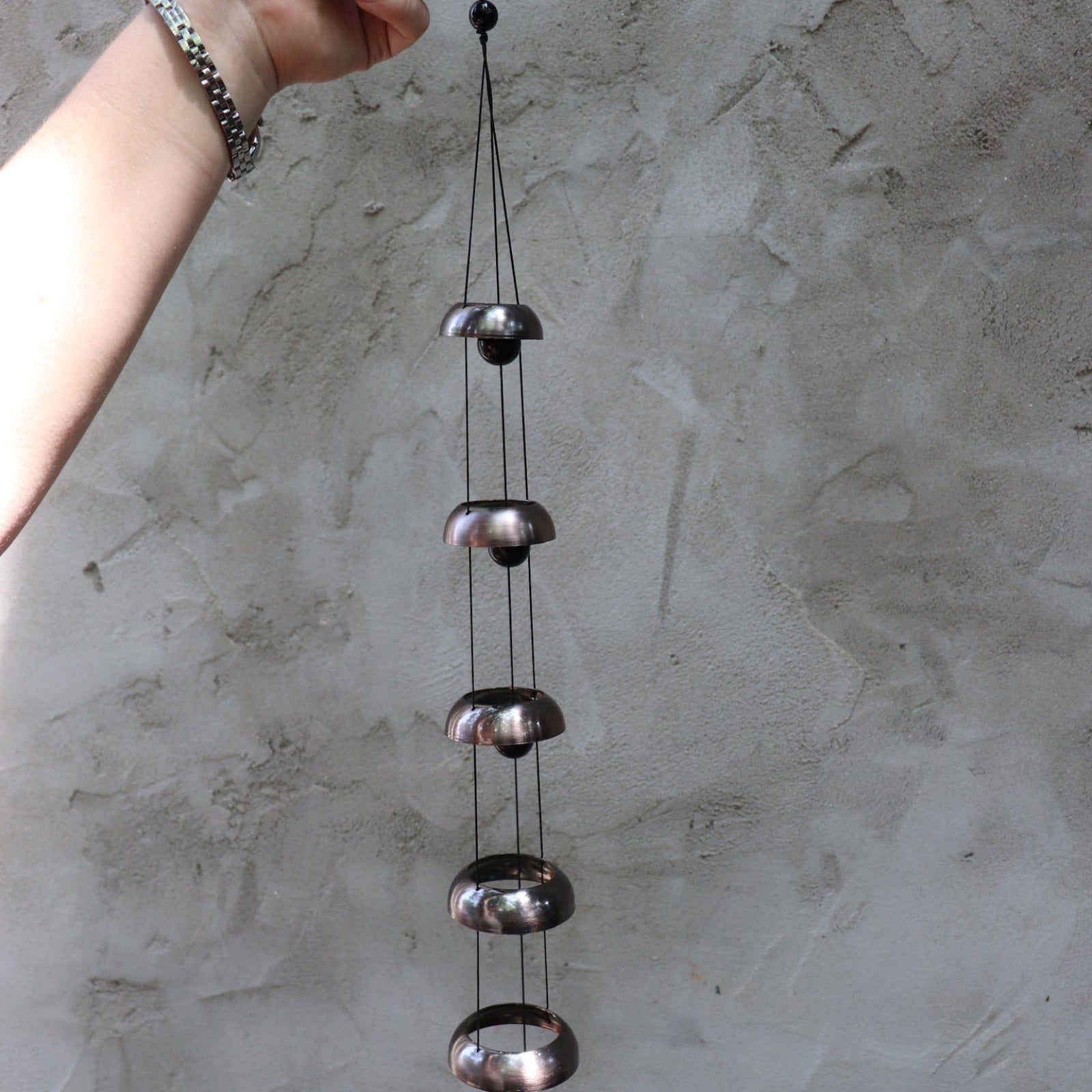 Someone holding the Copper Quintet Woodstock Temple Bells Wind Chime in front of a concrete wall.