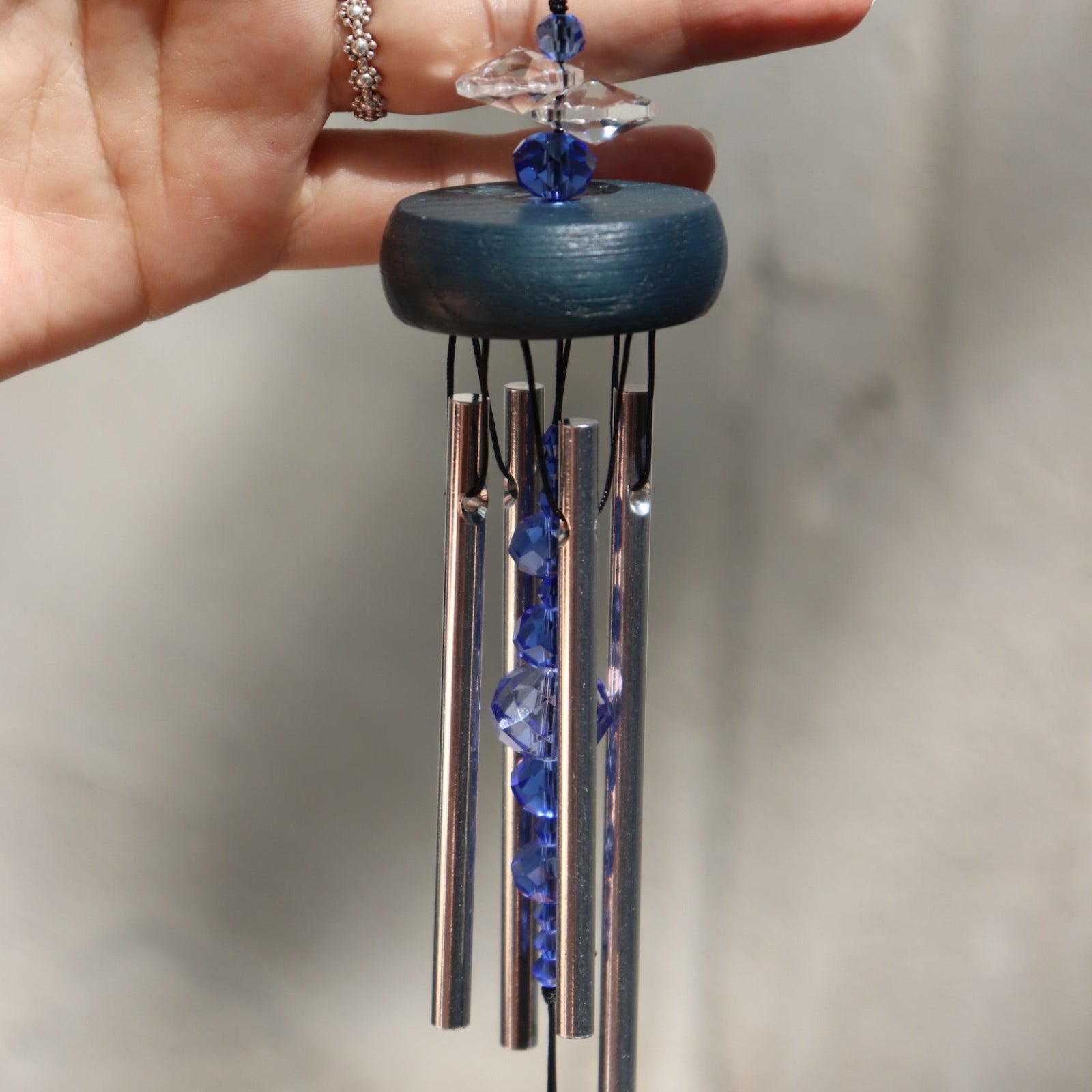 An up-close image of someone holding the Sapphire Gem Drop Wind Chime in front of a white cement wall.