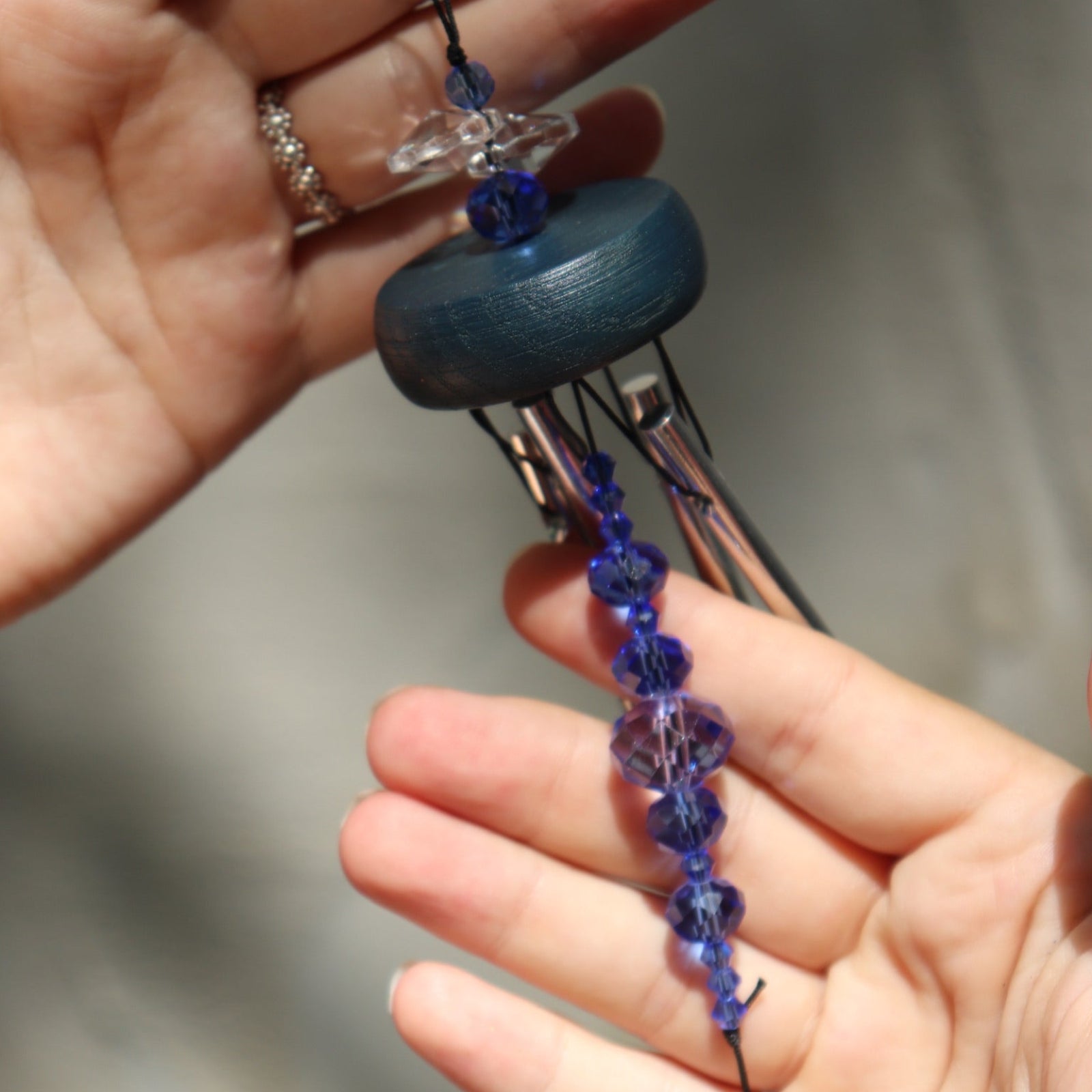 An up-close image of someone holding the Sapphire Gem Drop Wind Chime in front of a white cement wall.