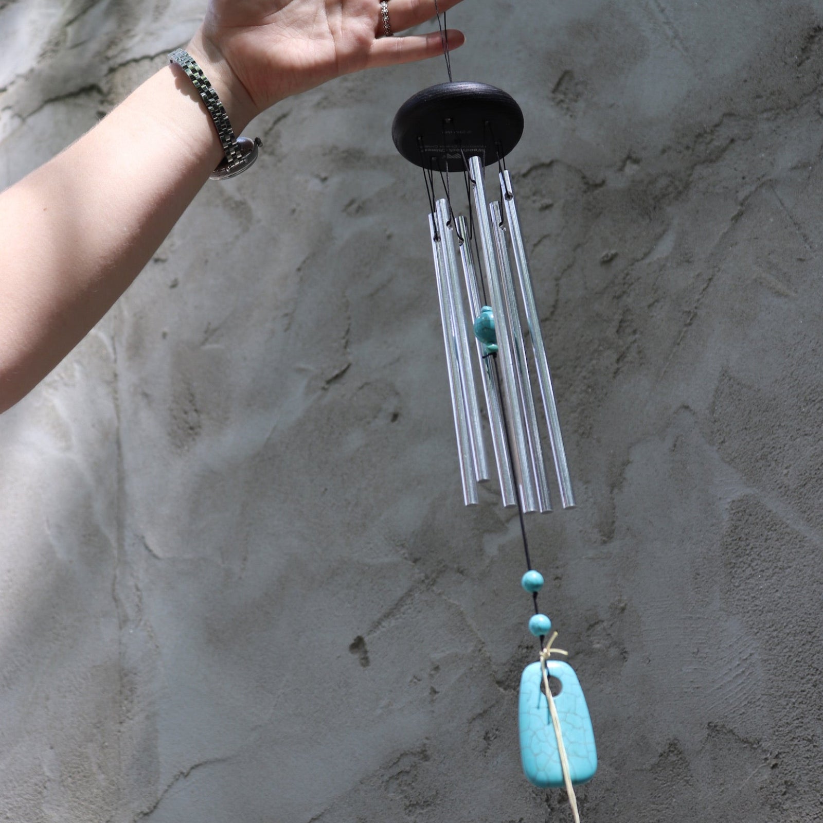 Someone holding the Turquoise Woodstock Chakra Wind Chime in front of a white wall.