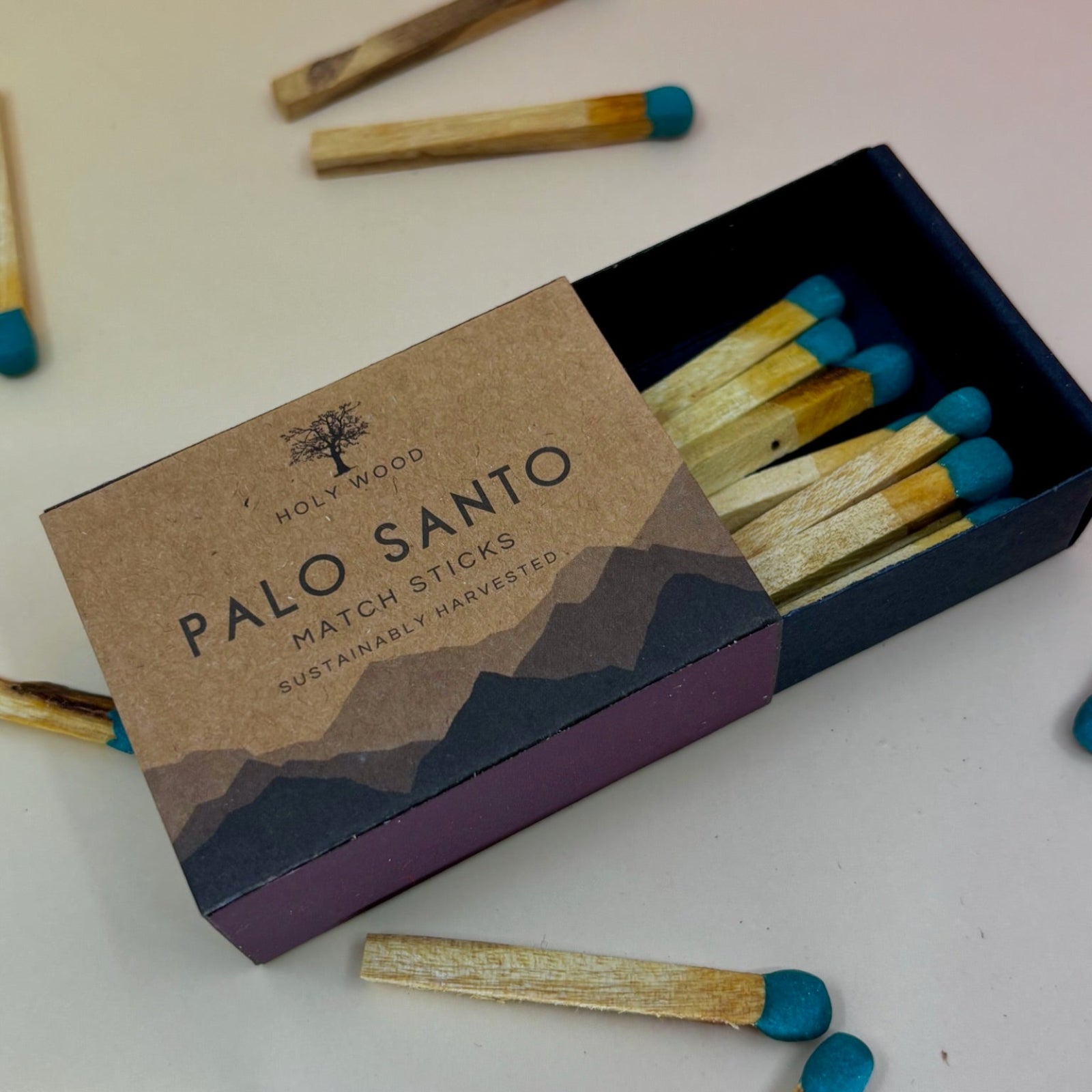 The Palo Santo Matchstick box on a pale pink and yellow background with matchsticks surrounding it.