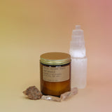 The Golden Coast - 7.2 oz Soy Wax Candle in front of a selenite tower surrounded by other crystals all on a pink and yellow background.