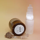 The Golden Coast - 7.2 oz Soy Wax Candle in front of a selenite tower surrounded by other crystals all on a pink and yellow background.