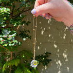 The Aquarius Zodiac Car Charm Suncatcher being held in front of a cream-colored brick wall with greenery.