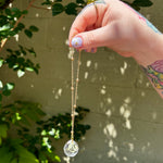 The Capricorn Zodiac Car Charm Suncatcher being held in front of a cream-colored brick wall with greenery.