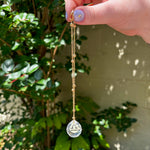 The Libra Zodiac Car Charm Suncatcher being held in front of a cream-colored brick wall with greenery.