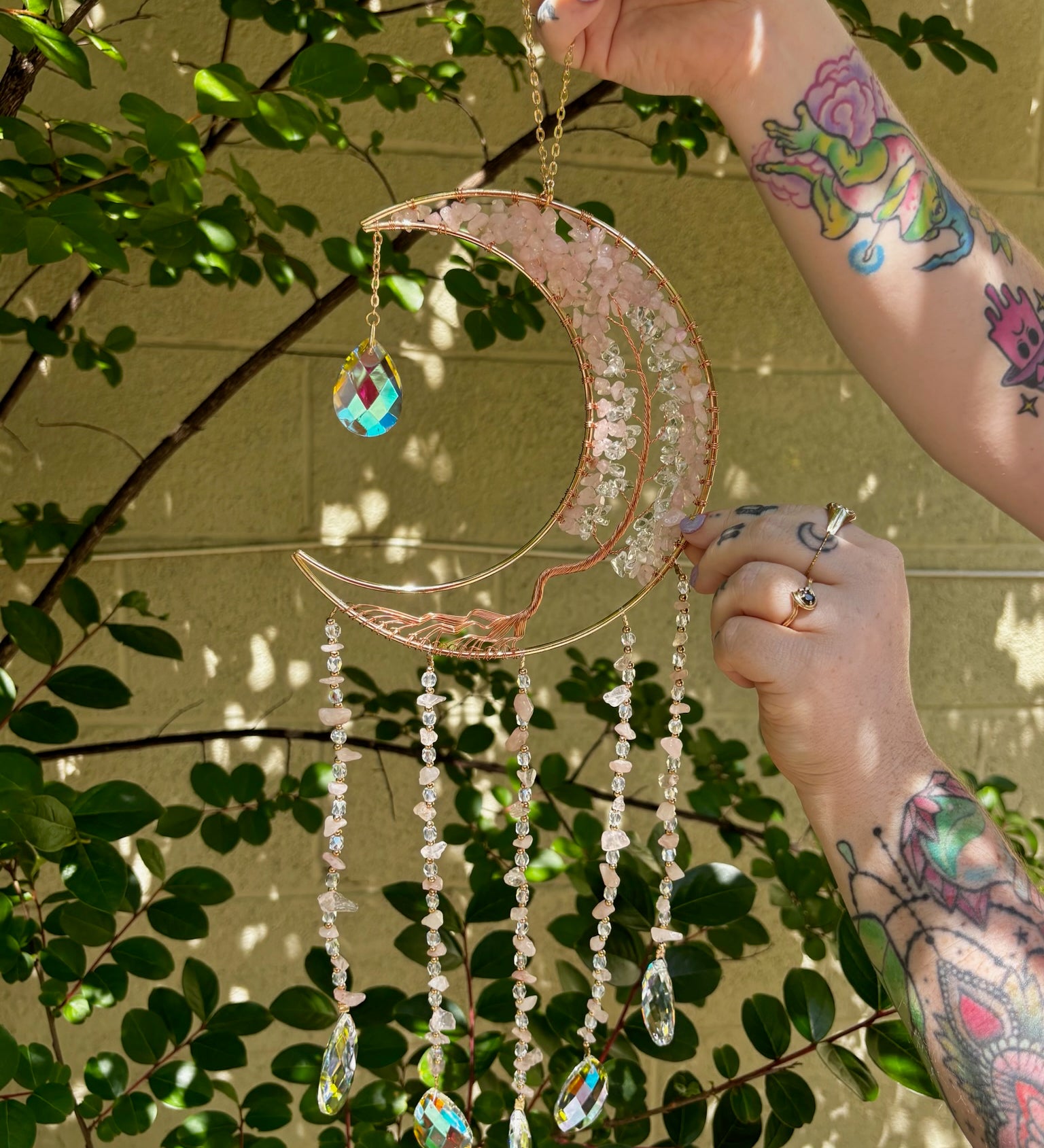 Someone holding the rose quartz Moon Crystal Gemstone Suncatcher in front of a cream-colored brick wall with greenery.