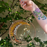 The Abundance Tree of Wealth Citrine Suncatcher being held in front of a cream-colored brick wall with greenery.