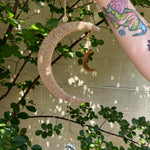 The Boho Woven Moon Suncatcher - Rustic Window Decor being held by someone in front of a cream-colored brick wall with greenery behind it.