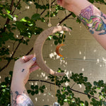 The Boho Woven Moon Suncatcher - Rustic Window Decor being held by someone in front of a cream-colored brick wall with greenery behind it.