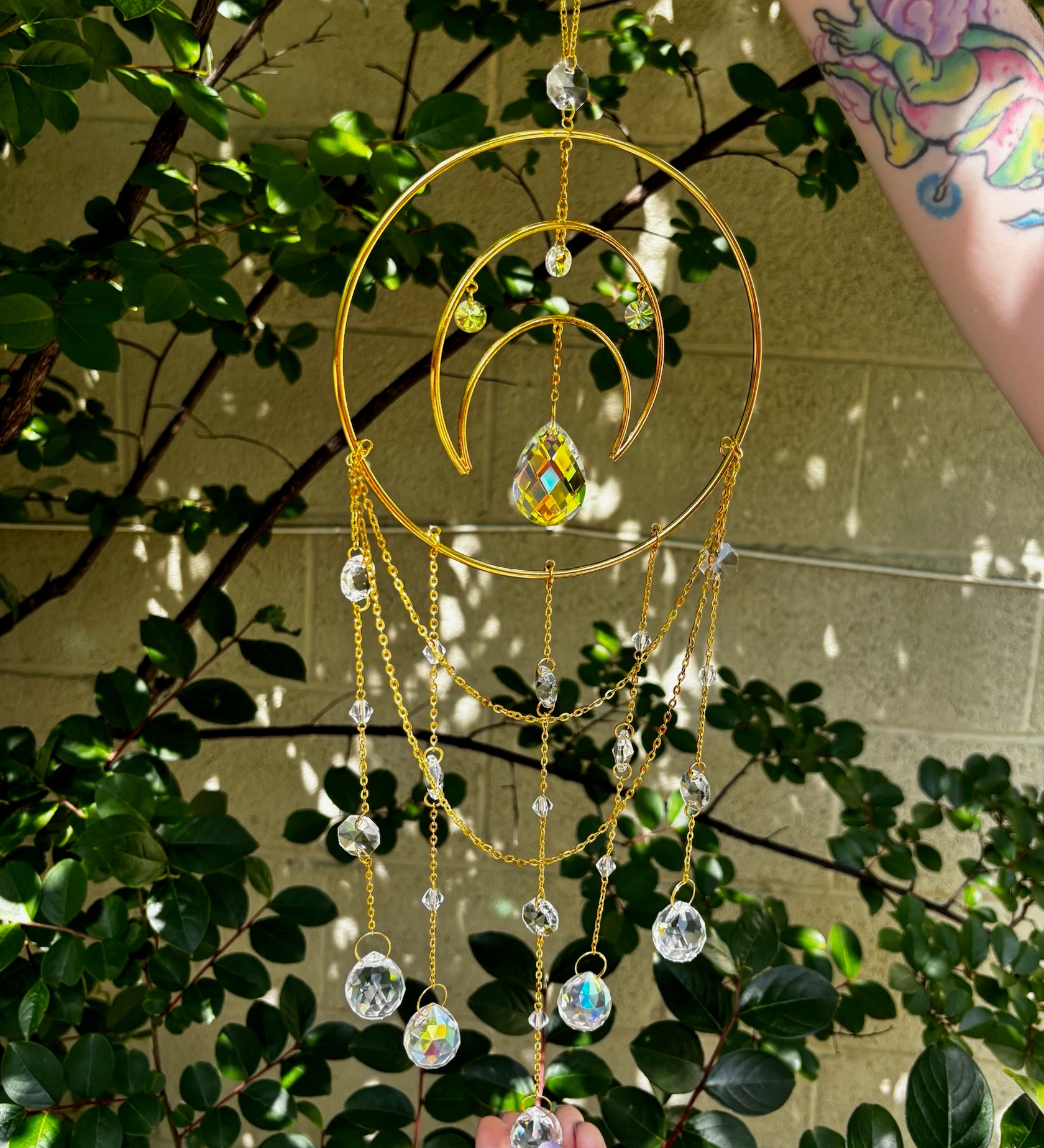 Someone holding the Large Celestial Suncatcher in front of a cream-colored brick wall with greenery behind it.