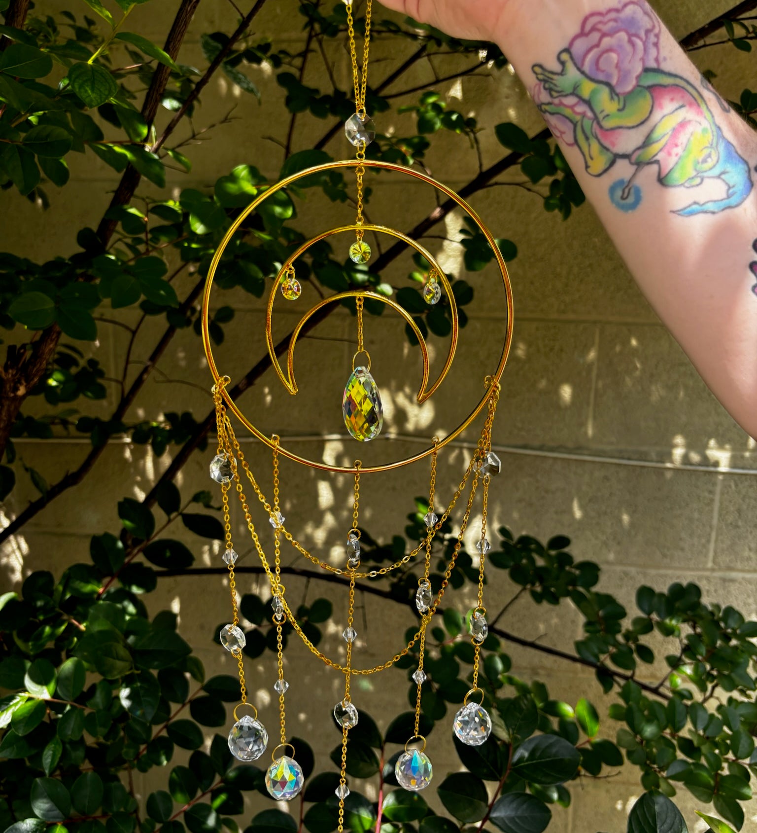 Someone holding the Large Celestial Suncatcher in front of a cream-colored brick wall with greenery behind it.