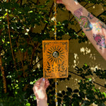 Someone holding the The Sun Wooden Tarot Suncatcher in front of a cream-colored brick wall with greenery.