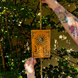 Someone holding the The Sun Wooden Tarot Suncatcher in front of a cream-colored brick wall with greenery.
