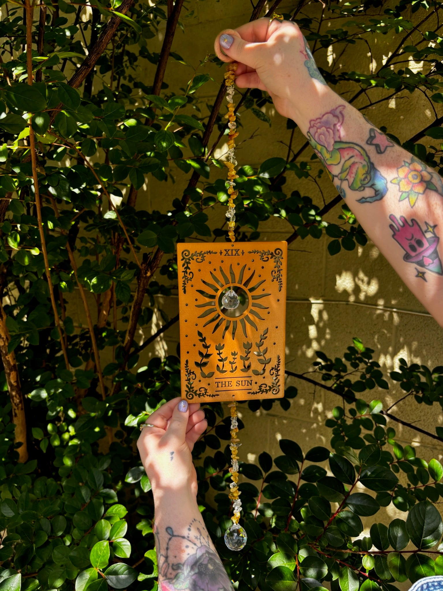 Someone holding the The Sun Wooden Tarot Suncatcher in front of a cream-colored brick wall with greenery.