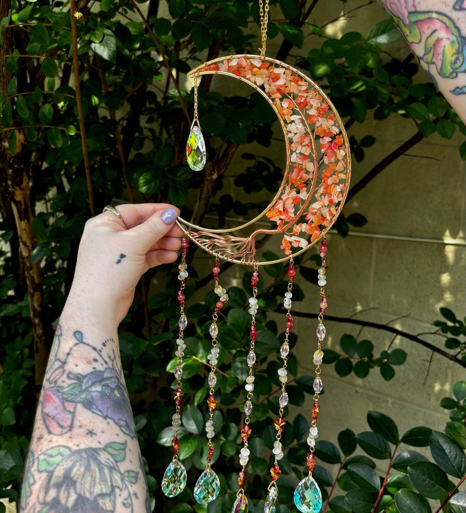 Someone holding the red agate Moon Crystal Gemstone Suncatcher in front of a cream-colored brick wall with greenery.