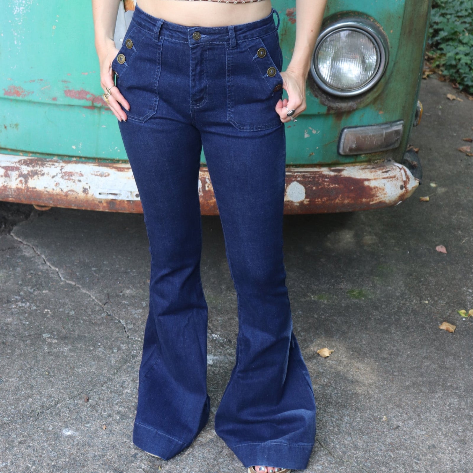 A woman wearing the Dark Denim Western Flared Jeans with Side Buttons with an orange, red, and brown crochet top all in front of a vintage Volkswagen bus.