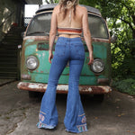 A woman wearing the Midtone Embroidered Denim Bell Bottoms and a red, orange, and brown crochet top in front of a vintage Volkswagen bus.