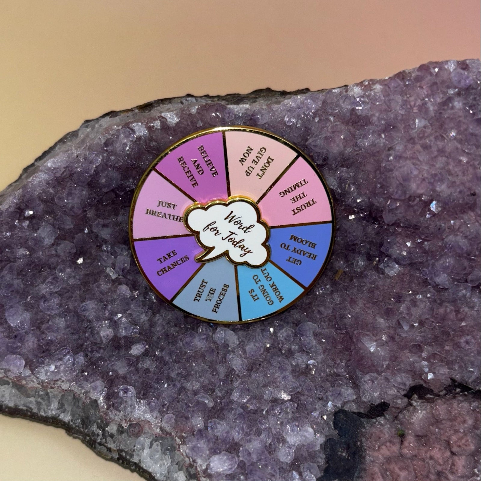 The Word for Today Enamel Pin on an amethyst piece all on a pink and yellow background.