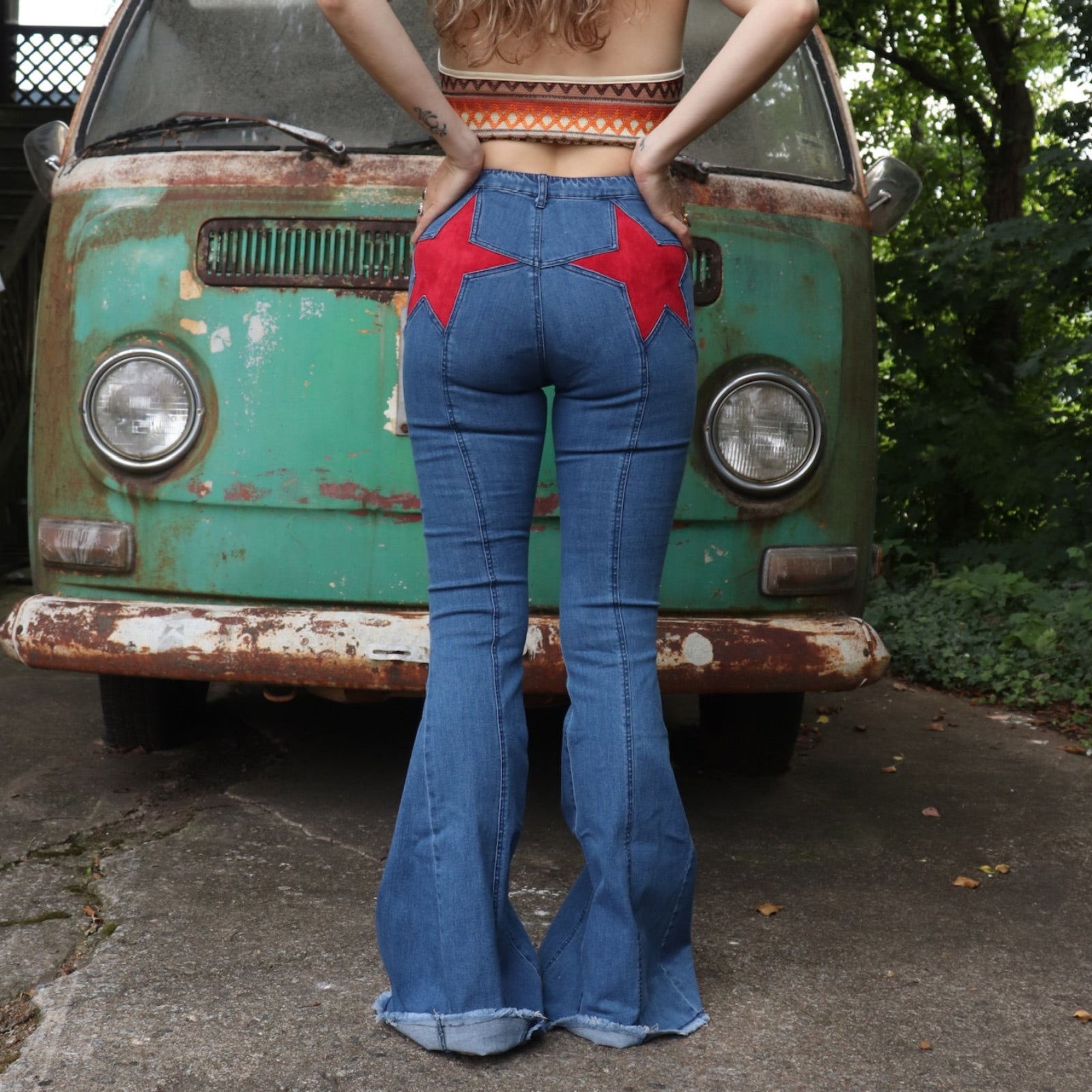 A woman wearing the Western Red Suede Stars Flared Denim Jeans with a red, orange, and brown top all in front of a vintage Volkswagen van.
