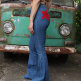 A woman wearing the Western Red Suede Stars Flared Denim Jeans with a red, orange, and brown top all in front of a vintage Volkswagen van.