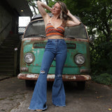 A woman wearing the Western Midtone Ripped and Fringed Stretchy Flared Jeans with a red, orange, and brown top all in front of a vintage Volkswagen bus.