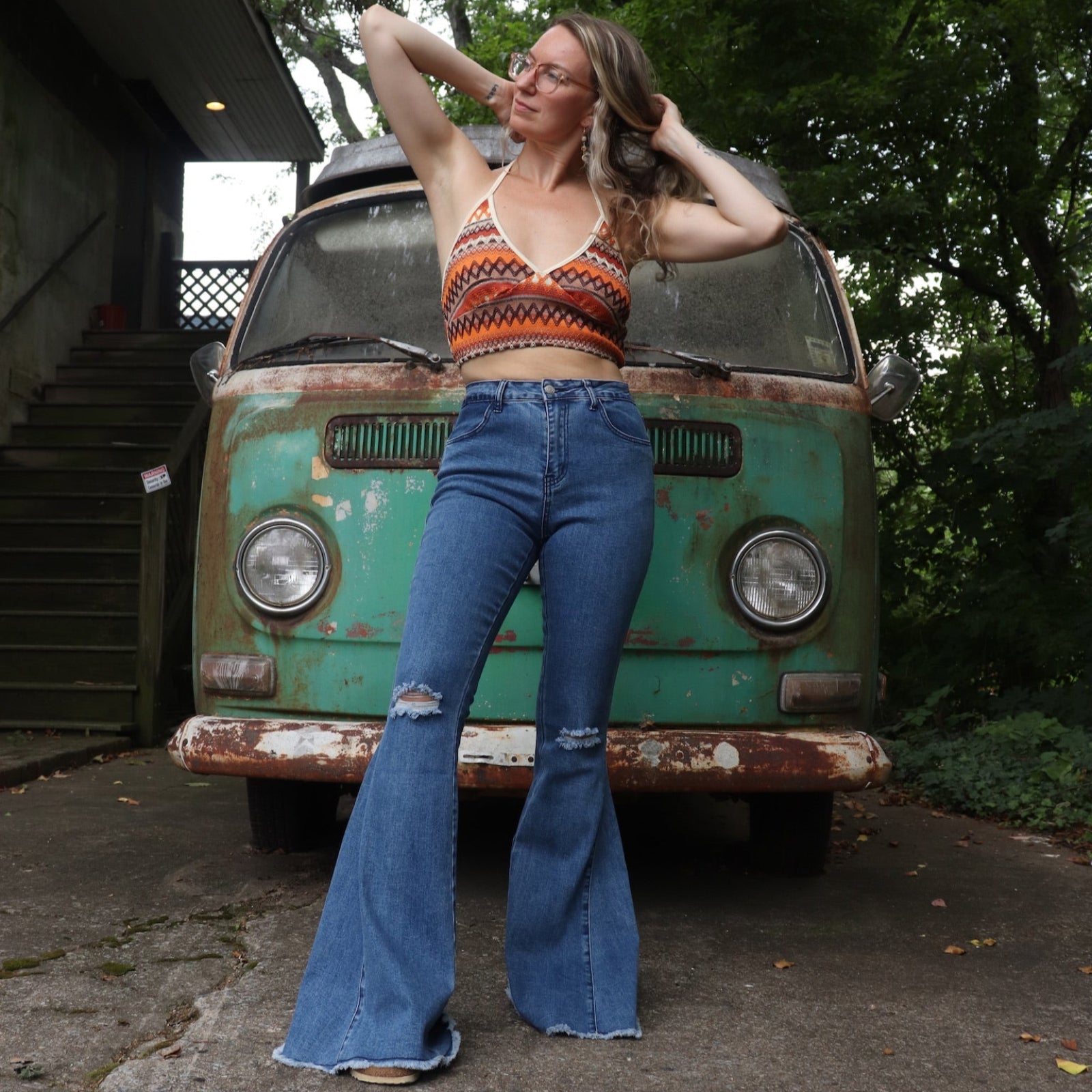 A woman wearing the Western Midtone Ripped and Fringed Stretchy Flared Jeans with a red, orange, and brown top all in front of a vintage Volkswagen bus.