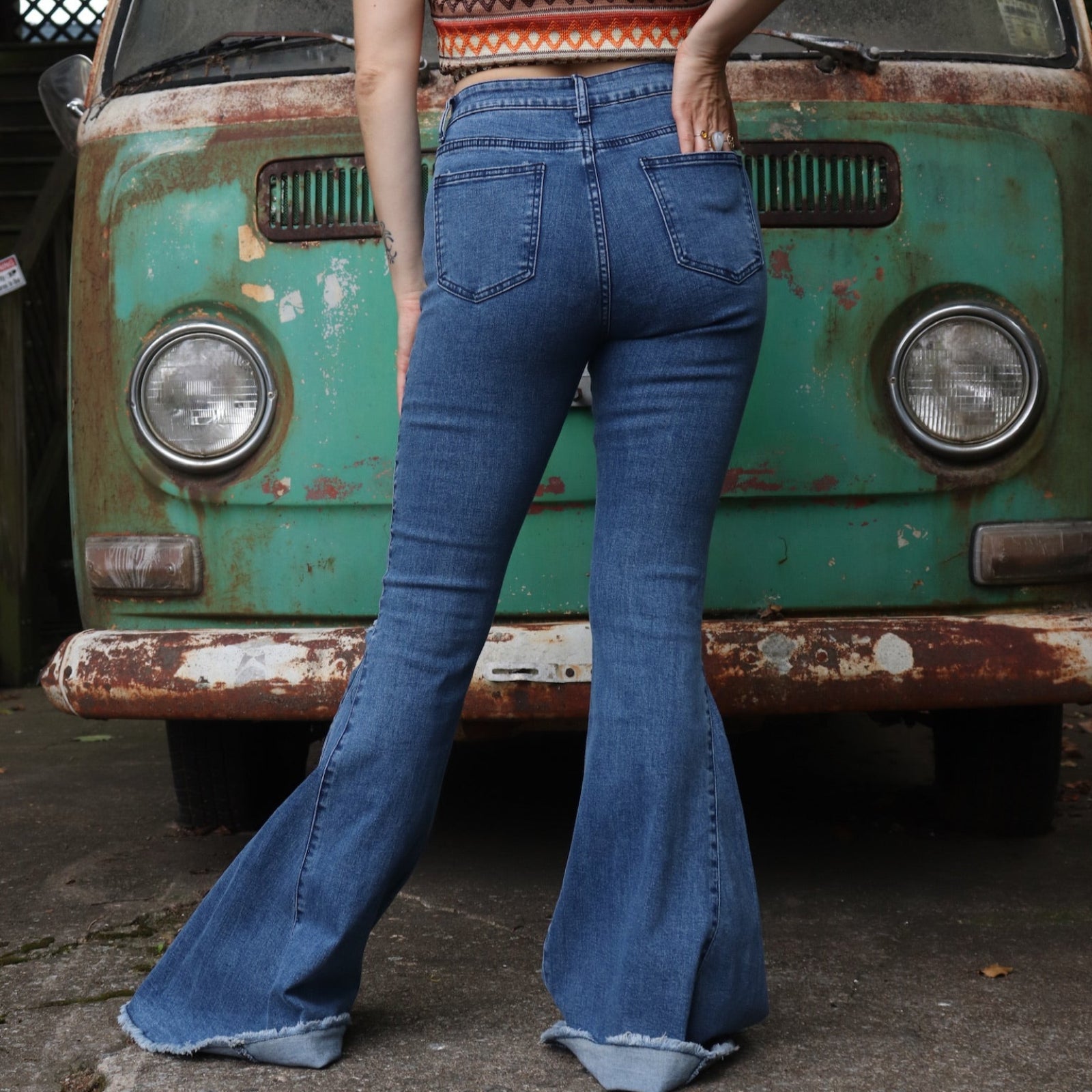 A woman wearing the Western Midtone Ripped and Fringed Stretchy Flared Jeans with a red, orange, and brown top all in front of a vintage Volkswagen bus.