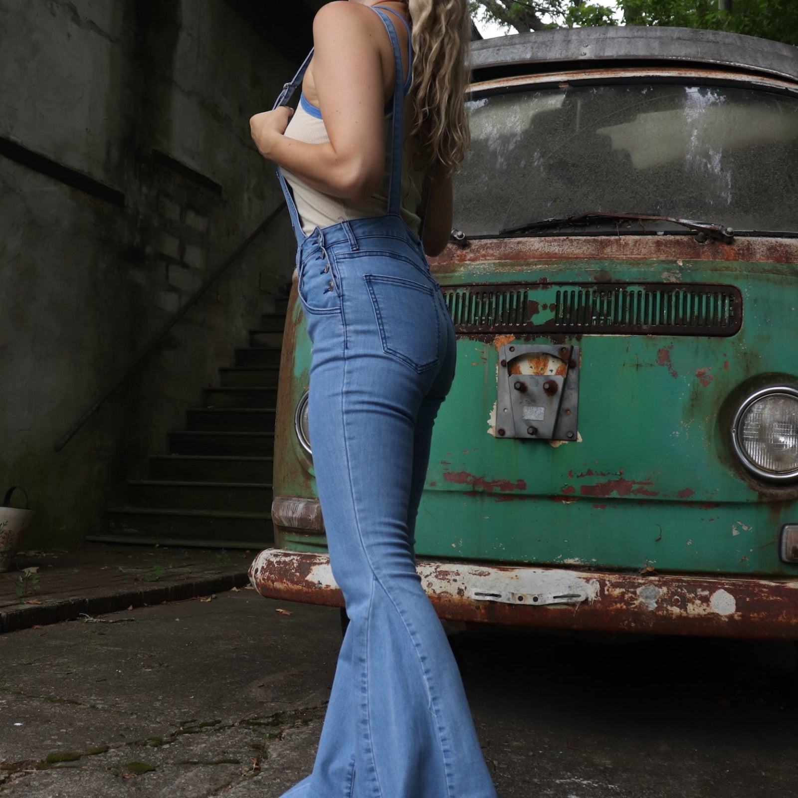 A woman wearing the light denim Bell Bottom Overalls with a cream tank all in front of a vintage Volkswagen bus.