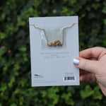 Someone holding the Taurus 14k Gold Dipped Nickel-Free Zodiac Necklace on its card in front of greenery showing off the back.