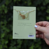 Someone holding the Virgo 14k Gold Dipped Nickel-Free Zodiac Necklace on its card in front of greenery showing off the back.