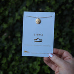 Someone holding the Libra 14k Gold Dipped Nickel-Free Zodiac Necklace on its card in front of greenery.
