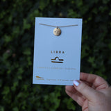Someone holding the Libra 14k Gold Dipped Nickel-Free Zodiac Necklace on its card in front of greenery.