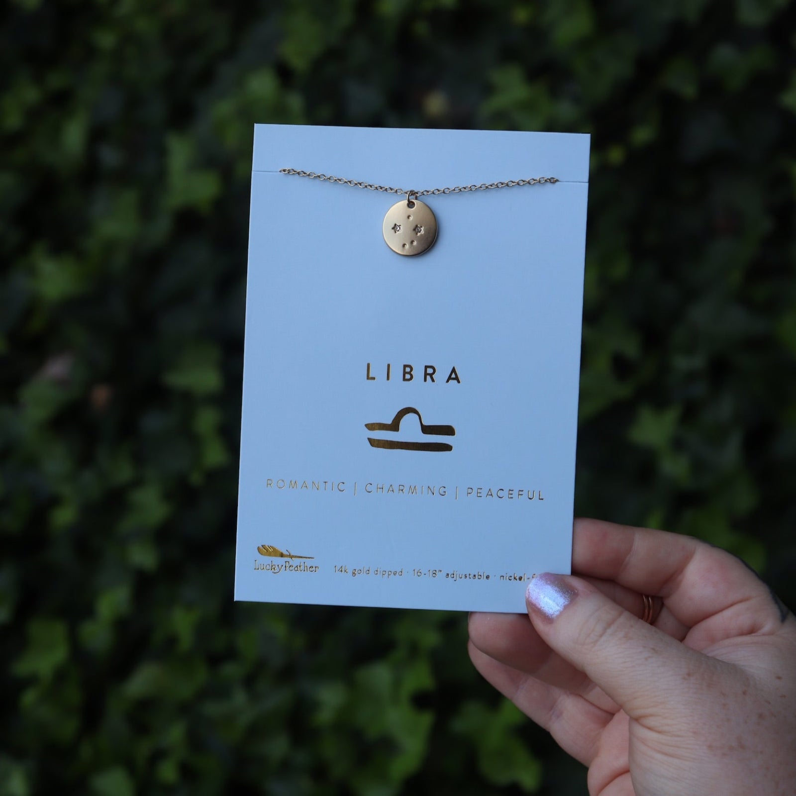 Someone holding the Libra 14k Gold Dipped Nickel-Free Zodiac Necklace on its card in front of greenery.