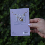 Someone holding the Pisces 14k Gold Dipped Nickel-Free Zodiac Necklace on its card in front of greenery showing off the back.