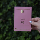 Someone holding the Leo 14k Gold Dipped Nickel-Free Zodiac Necklace on its card in front of greenery.