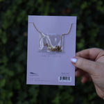 Someone holding the Cancer 14k Gold Dipped Nickel-Free Zodiac Necklace on its card in front of greenery showing off the back.