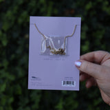 Someone holding the Cancer 14k Gold Dipped Nickel-Free Zodiac Necklace on its card in front of greenery showing off the back.