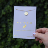 Someone holding the Sagittarius 14k Gold Dipped Nickel-Free Zodiac Necklace on its card in front of greenery.