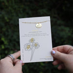 Someone holding the daisy 14k Gold Dipped Nickel-Free Birth Month Flower Necklace in front of greenery.