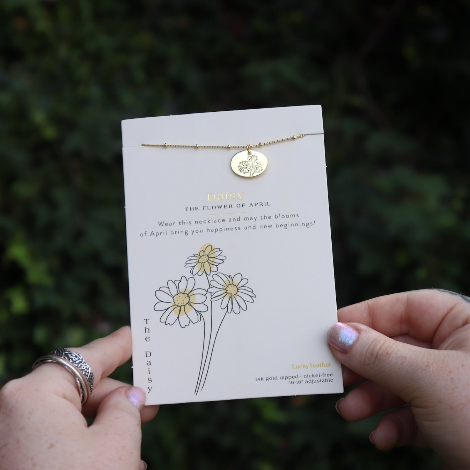 Someone holding the daisy 14k Gold Dipped Nickel-Free Birth Month Flower Necklace in front of greenery.