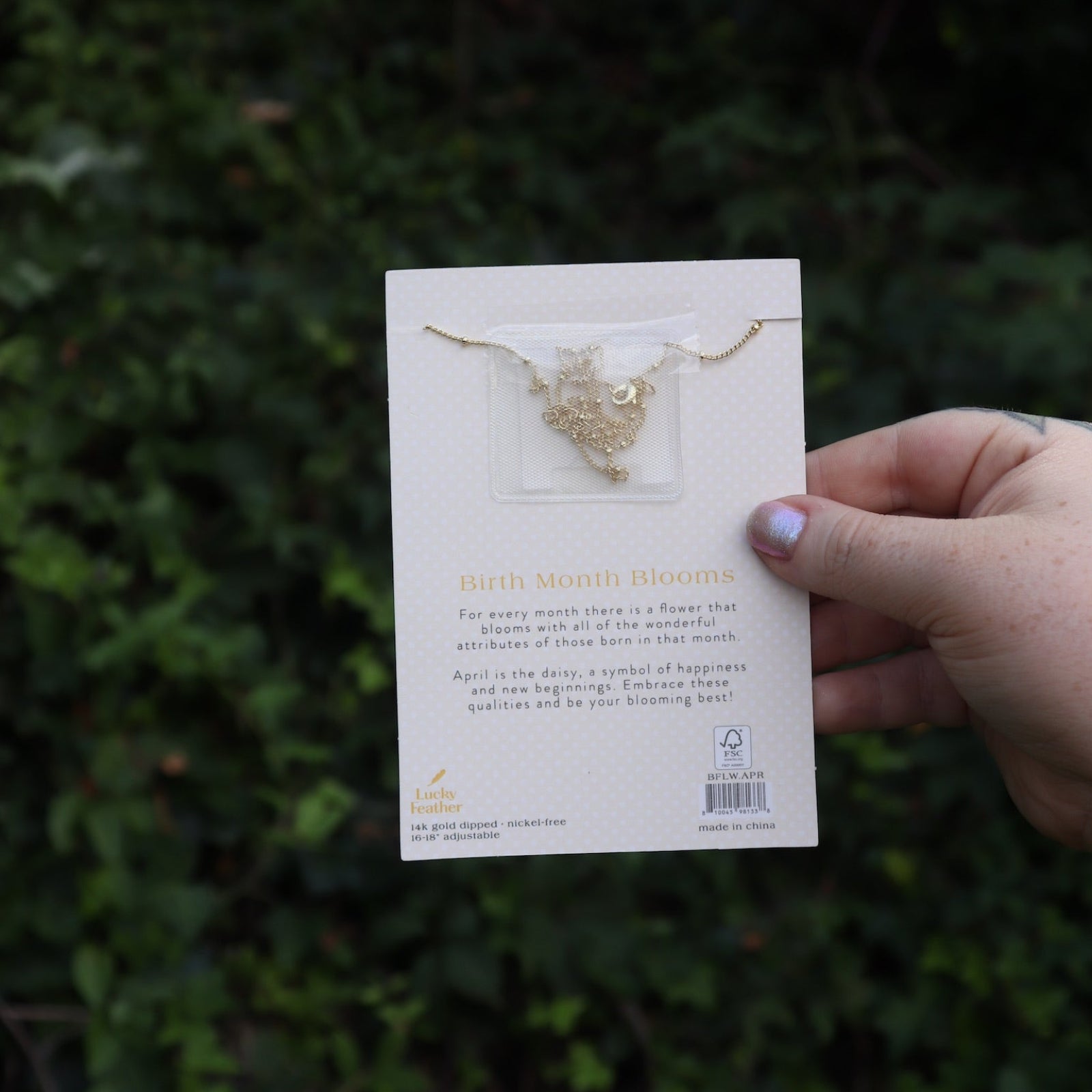 Someone holding the daisy 14k Gold Dipped Nickel-Free Birth Month Flower Necklace in front of greenery showing off the back.