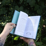 Someone holding the Spell Jars for the Modern Witch Book in front of greenery, showing off the pages.