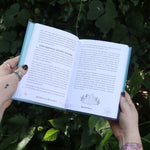 Someone holding the Spell Jars for the Modern Witch Book in front of greenery, showing off the pages.