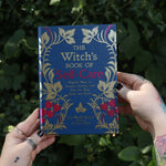 Someone holding the Witch's Book of Self-Care in front of greenery.