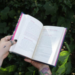 Someone holding the Witch's Book of Self-Care in front of greenery, showing off the pages.