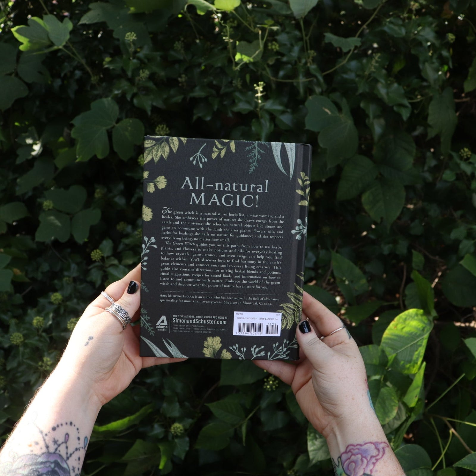 Someone holding the Green Witch by Arin Murphy-Hiscock in front of greenery, showing off the back.