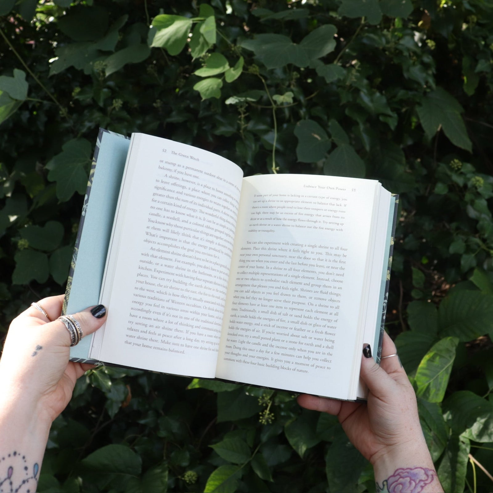 Someone holding the Green Witch by Arin Murphy-Hiscock open in front of greenery, showing off the pages.