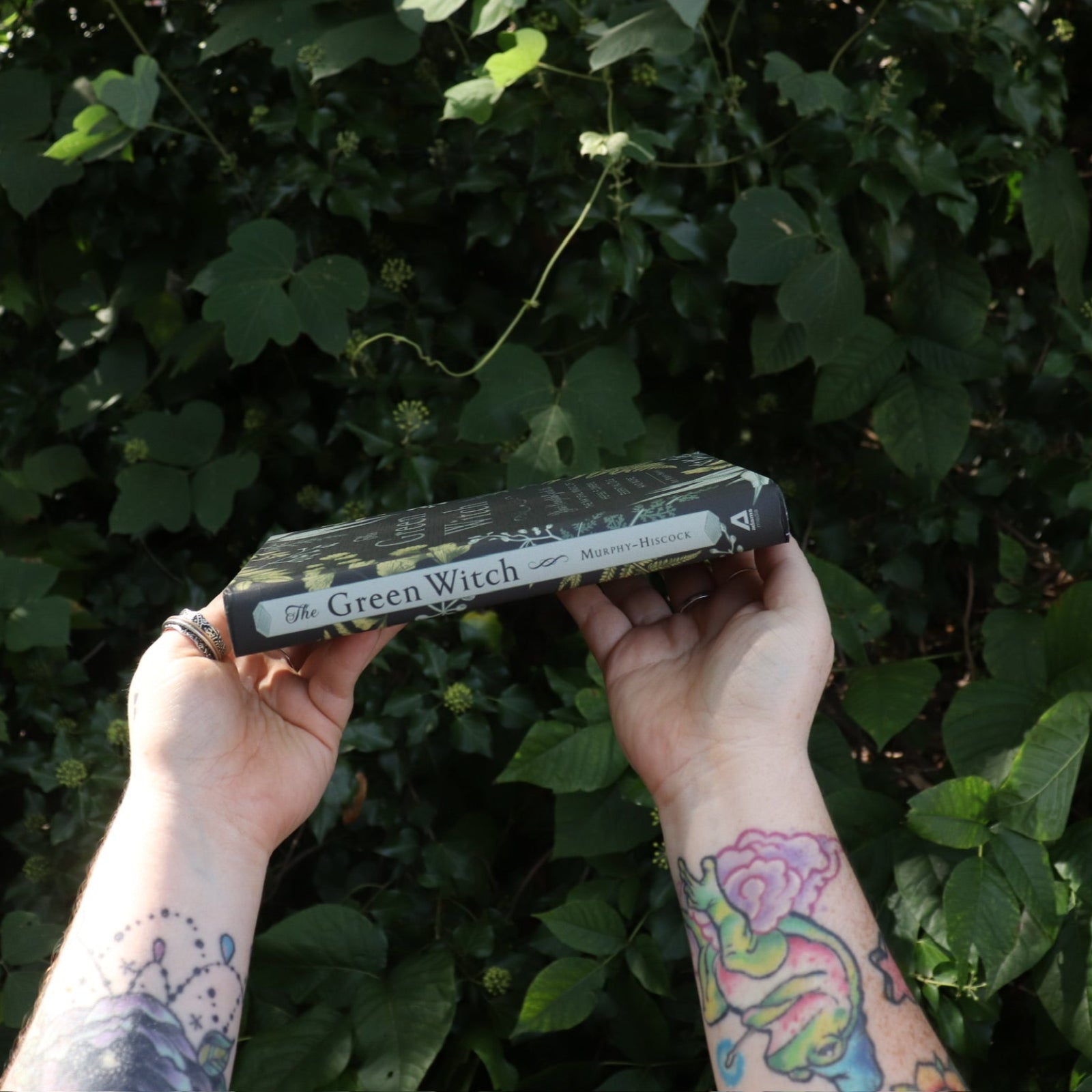 Someone holding the Green Witch by Arin Murphy-Hiscock in front of greenery, showing off the spine of the book.